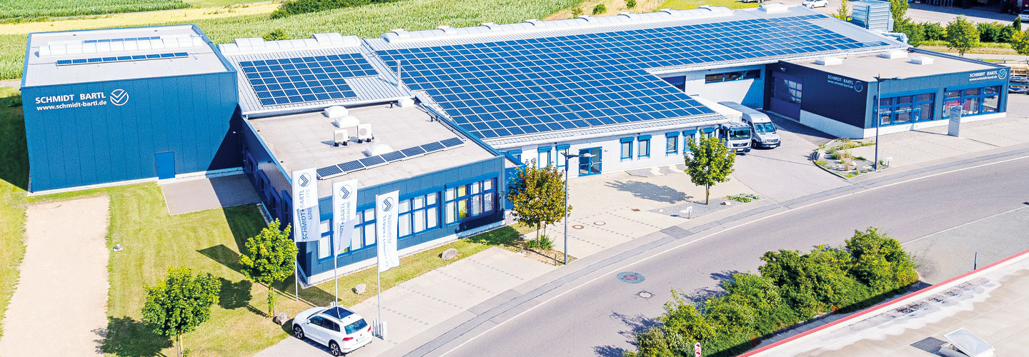 Das Unternehmensgebäude ist von einer grünen Rasenfläche umgeben, während eine Straße daran vorbeiführt. Mehrere moderne Werkhallen wurden kürzlich an das Hauptgebäude angebaut.