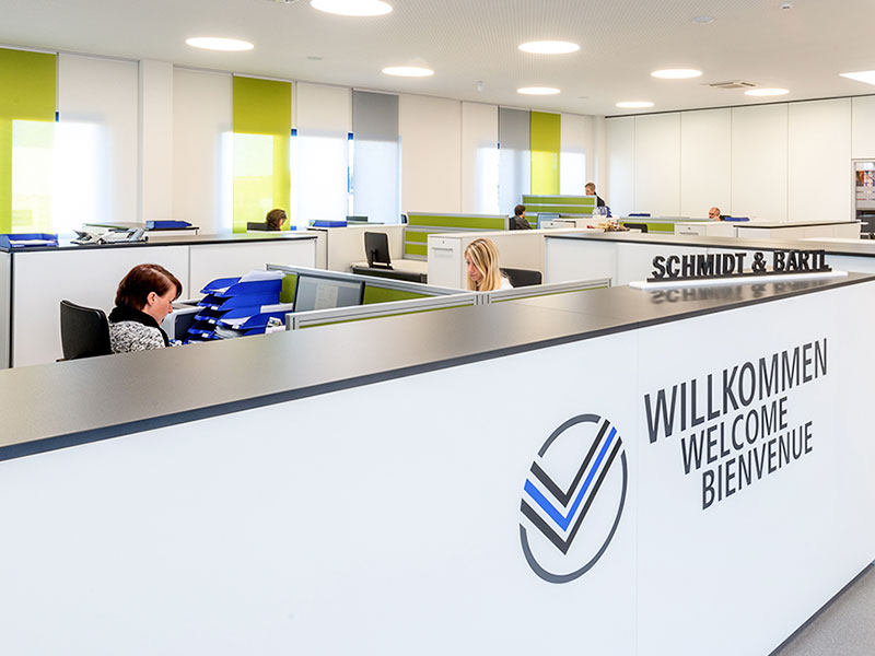 Blick in das Büro von Schmidt+Bartl: Im Vordergrund steht ein langer weißer Tresen. Dahinter sind mehrere kleine Arbeitskojen zu sehen, von denen einige von Mitarbeitern besetzt sind, die konzentriert arbeiten.