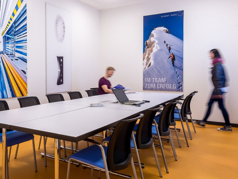 Ein langer Konferenztisch steht in einem weißen Raum. An den Wänden hängen mehrere farbige, lange Banner. In der Mitte des Tisches liegt ein aufgeklapptes Laptop. Am Ende des Tisches sitzt ein Mann, der dem herantretenden Kunden entgegenblickt.